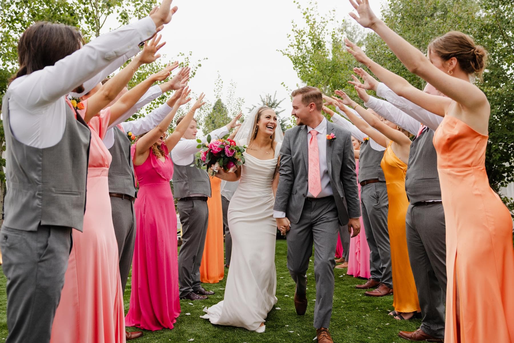 Bride and groom walking hand in hand through a joyful tunnel of bridesmaids in coral and orange dresses, Colorado wedding