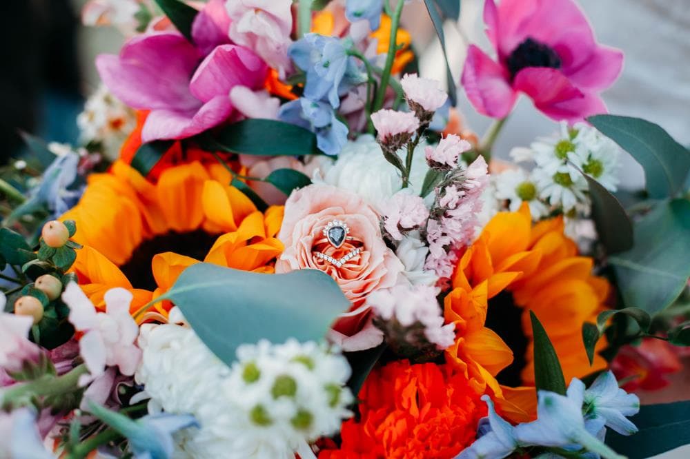 Colorful wedding bouquet of sunflowers, pink anemones and blue delphinium cradling the engagement ring, Colorado wedding